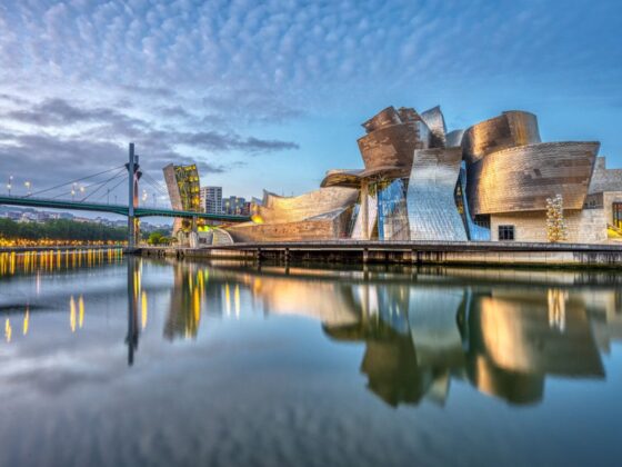 The Guggenheim Museum Bilbao's sculptural metallic façade reflects in calm river water at dusk, with a bridge to the left and city lights in the background.