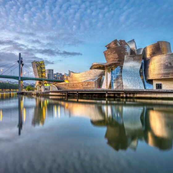 The Guggenheim Museum Bilbao's sculptural metallic façade reflects in calm river water at dusk, with a bridge to the left and city lights in the background.