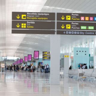 Airport terminal corridor with overhead signs directing to baggage claim, exits, gates, and Sky Centre in multiple languages.