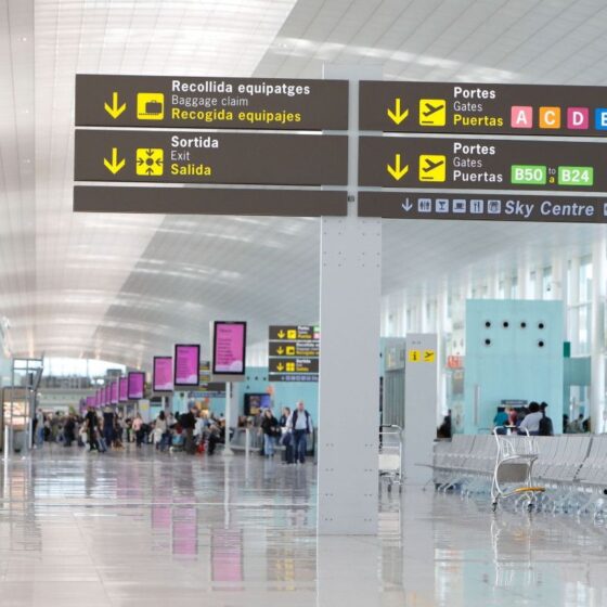 Airport terminal corridor with overhead signs directing to baggage claim, exits, gates, and Sky Centre in multiple languages.