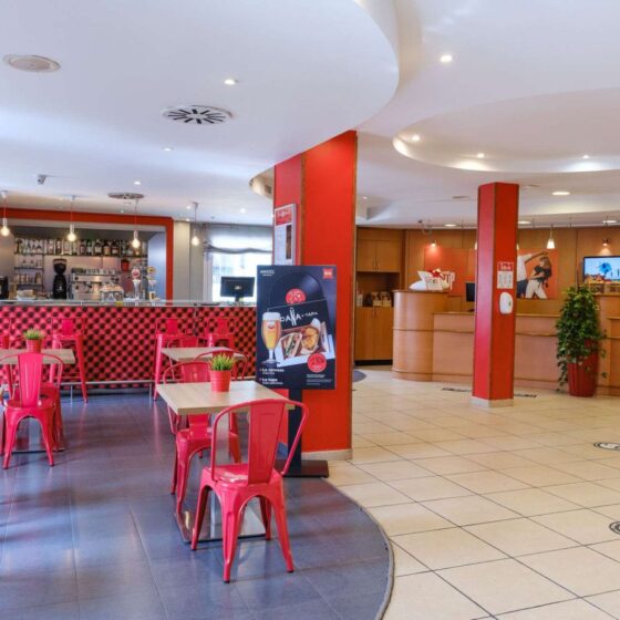 Cafe interior with red metal chairs and light tables, curved white ceiling, and a red-and-black checkered bar front behind the counter.