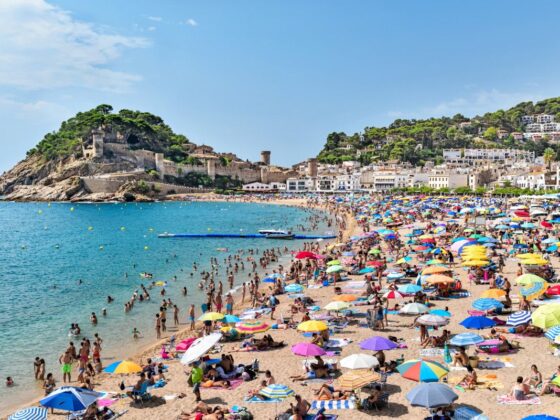 Crowded sunny beach along a rocky coastline, with colorful umbrellas, sunbathers, and clear blue water.