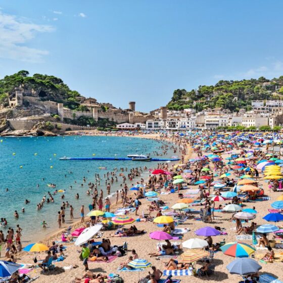 Crowded sunny beach along a rocky coastline, with colorful umbrellas, sunbathers, and clear blue water.