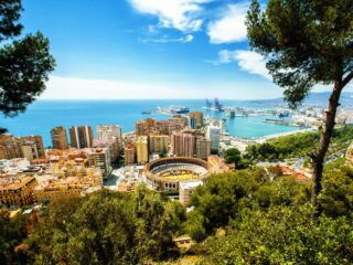 Panoramic view of a coastal city with a circular arena, colorful buildings, and a blue sea on the horizon.