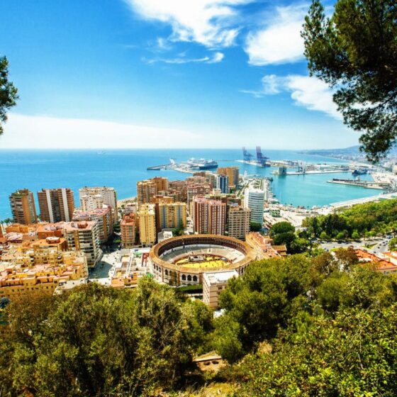 Panoramic view of a coastal city with a circular arena, colorful buildings, and a blue sea on the horizon.