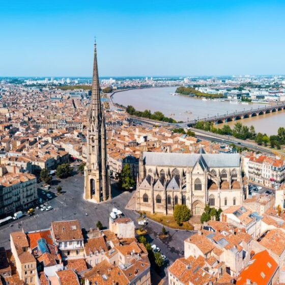 Aerial view of a European city with a tall Gothic cathedral, red-tile roofs, and a river with a bridge in the background.