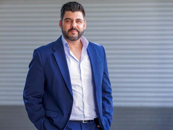Man in a navy suit with a light striped shirt posing outdoors against a gray corrugated wall.