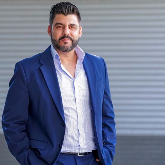 Man in a navy suit with a light striped shirt posing outdoors against a gray corrugated wall.