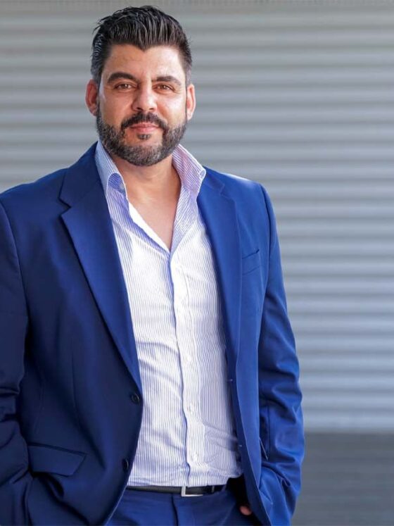 Man in a navy suit with a light striped shirt posing outdoors against a gray corrugated wall.