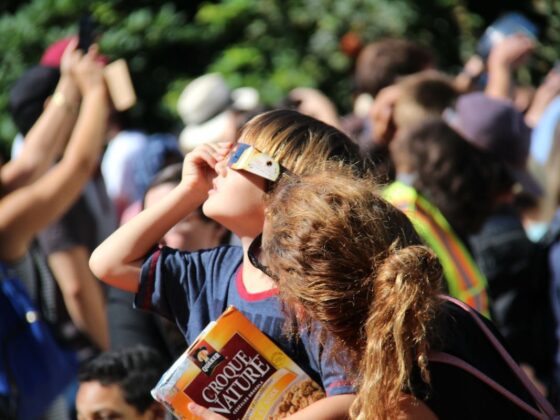 Young boy wearing makeshift sunglasses shields his eyes in a crowded outdoor scene, clutching a box of cereal/snack.