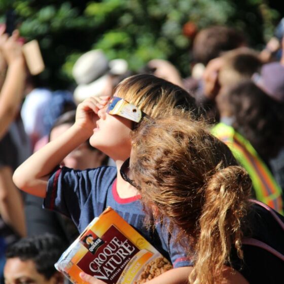 Young boy wearing makeshift sunglasses shields his eyes in a crowded outdoor scene, clutching a box of cereal/snack.