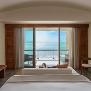 Hotel room with a balcony and a panoramic ocean view, seen from the foot of a neatly made bed with a sofa and coffee table nearby, plus wood furniture. Check the sea through large sliding doors.