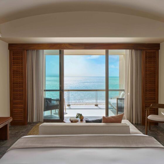 Hotel room with a balcony and a panoramic ocean view, seen from the foot of a neatly made bed with a sofa and coffee table nearby, plus wood furniture. Check the sea through large sliding doors.