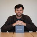 Smiling man sits at a table with hands folded, a gray box labeled 'HOTEL SPEAKER' in front of him on the table.