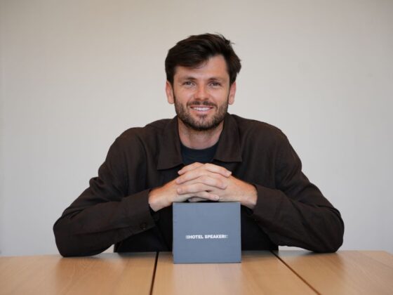 Smiling man sits at a table with hands folded, a gray box labeled 'HOTEL SPEAKER' in front of him on the table.