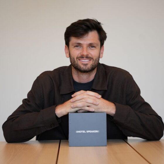 Smiling man sits at a table with hands folded, a gray box labeled 'HOTEL SPEAKER' in front of him on the table.