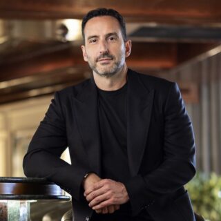 Portrait of a man in a black blazer standing and leaning on a marble railing in a warmly lit indoor space, looking at the camera.