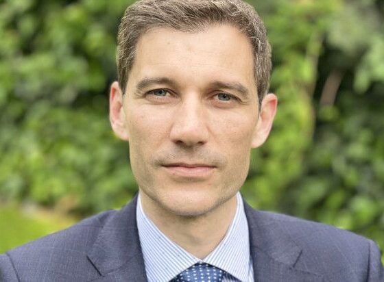 Head-and-shoulders portrait of a man in a navy suit and light blue shirt, outdoors with green foliage in the background, neutral expression.