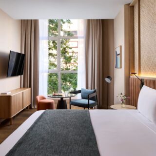 Hotel bedroom with large window, seating area, wall-mounted TV, and a king bed with white linens.
