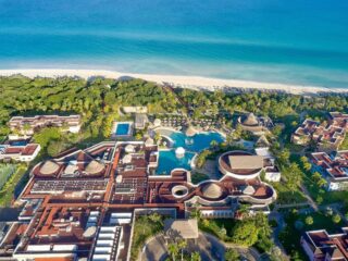 Aerial view of a tropical beachfront resort with multiple pools, palm trees, and red-roofed buildings along a white-sand shore.