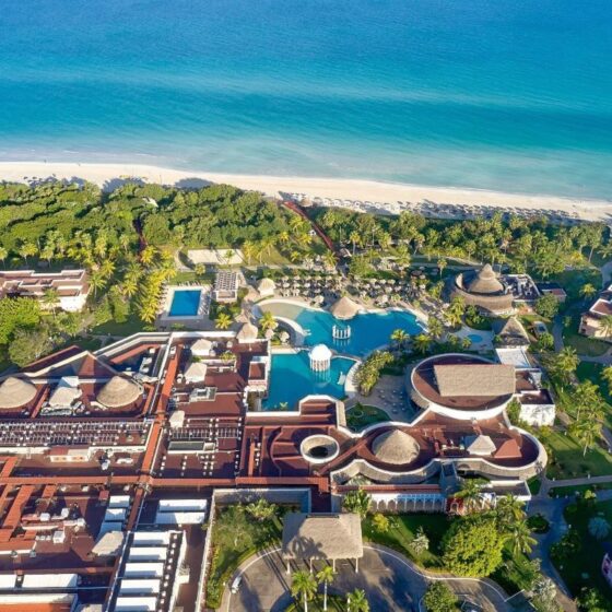 Aerial view of a tropical beachfront resort with multiple pools, palm trees, and red-roofed buildings along a white-sand shore.
