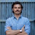 Man with dark hair and a mustache, wearing a light blue denim shirt and watch, stands with arms crossed before a metal fence.