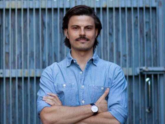 Man with dark hair and a mustache, wearing a light blue denim shirt and watch, stands with arms crossed before a metal fence.