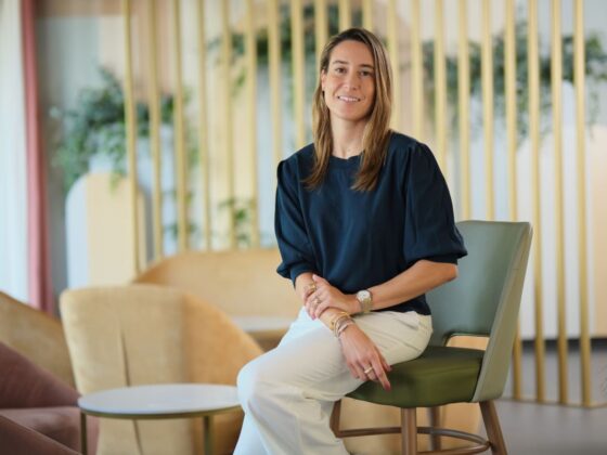 Woman seated on a green chair in a bright modern office lounge, smiling at the camera, wearing a navy blouse and white pants.