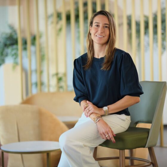 Woman seated on a green chair in a bright modern office lounge, smiling at the camera, wearing a navy blouse and white pants.