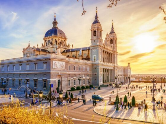 Sunlit cathedral with a blue dome and two tall towers, crowds on a wide plaza at sunset.