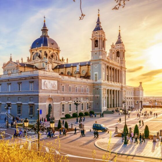 Sunlit cathedral with a blue dome and two tall towers, crowds on a wide plaza at sunset.