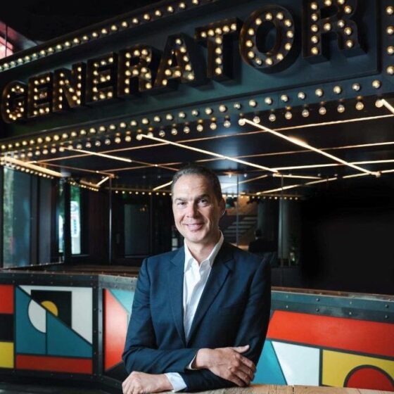 Smiling man in a navy suit stands at a wooden railing inside a venue with a bright, marquee-style sign reading 'GENERATOR' overhead.