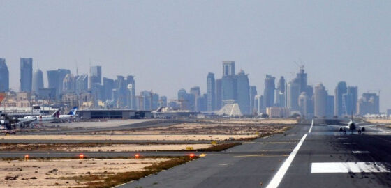 Runway extending toward a distant city skyline with parked planes to the left under a clear blue sky.