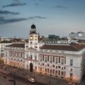 Wide view of a European plaza with a central clock tower atop a neoclassical building at dusk/light evening sky (blue).