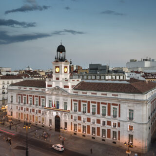 Wide view of a European plaza with a central clock tower atop a neoclassical building at dusk/light evening sky (blue).