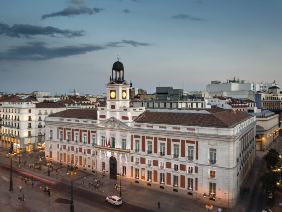 Wide view of a European plaza with a central clock tower atop a neoclassical building at dusk/light evening sky (blue).