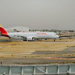 Iberia airplane on a taxiway at a sunny, dry airport with distant buildings in the background.