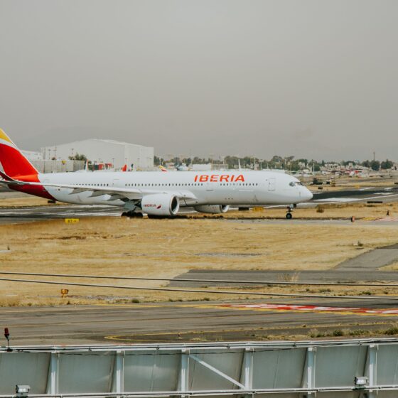Iberia airplane on a taxiway at a sunny, dry airport with distant buildings in the background.