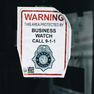 Warning sign on a glass window reading 'This area protected by business watch' and 'Call 9-1-1' with a police emblem and 'Lock Out Crime' note at the bottom, taped to the pane.