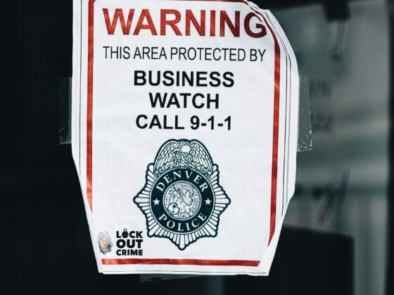 Warning sign on a glass window reading 'This area protected by business watch' and 'Call 9-1-1' with a police emblem and 'Lock Out Crime' note at the bottom, taped to the pane.