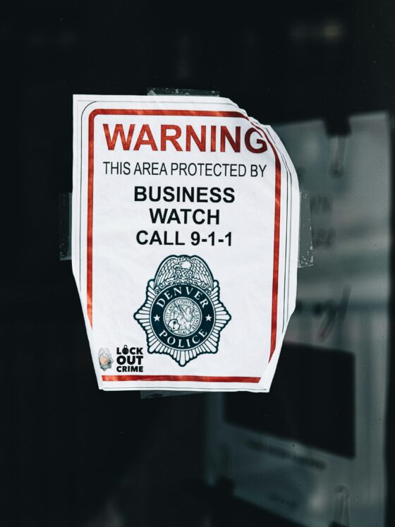 Warning sign on a glass window reading 'This area protected by business watch' and 'Call 9-1-1' with a police emblem and 'Lock Out Crime' note at the bottom, taped to the pane.