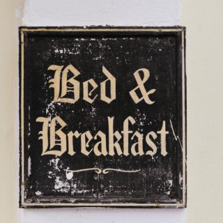Close-up of a weathered black sign with ornate gold Gothic letters mounted on a pale wall; lettering is chipped and faded in places.