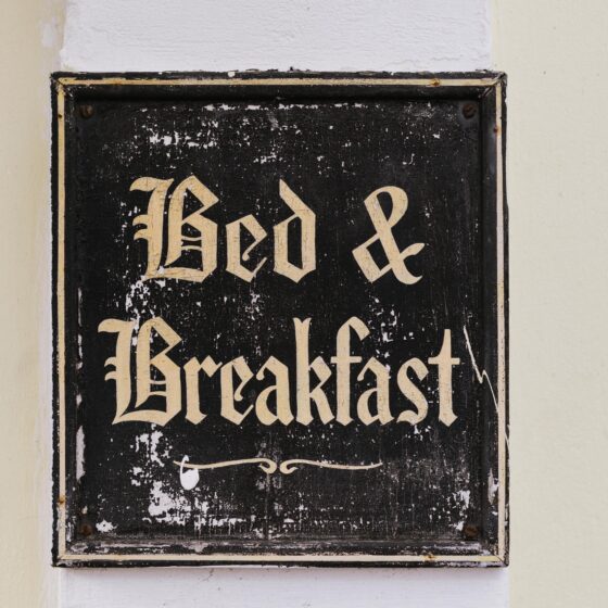 Close-up of a weathered black sign with ornate gold Gothic letters mounted on a pale wall; lettering is chipped and faded in places.