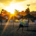 Man running barefoot on a sandy beach at sunset, with straw umbrellas and striped beach huts in the background and sun rays bursting through palm trees.