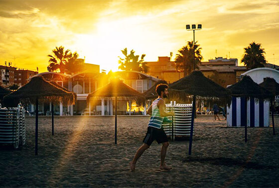 Man running barefoot on a sandy beach at sunset, with straw umbrellas and striped beach huts in the background and sun rays bursting through palm trees.
