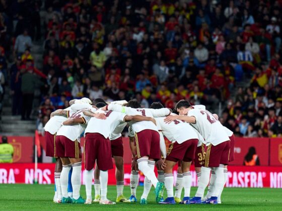 Soccer team in white jerseys huddling with arms around each other on the green field before kickoff, stadium crowd in the background.