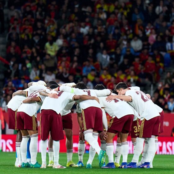 Soccer team in white jerseys huddling with arms around each other on the green field before kickoff, stadium crowd in the background.