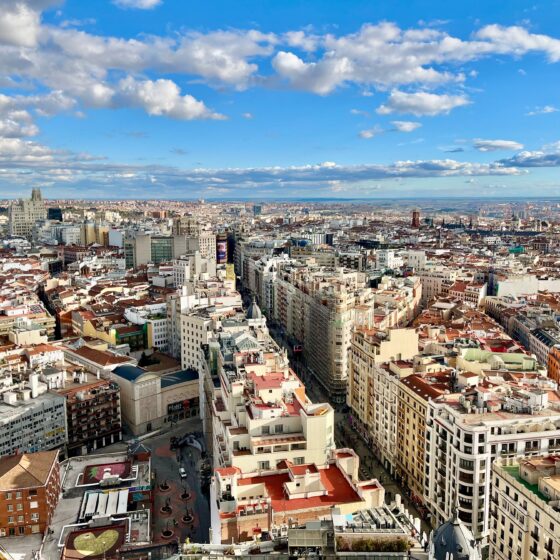 Aerial view of a dense city with red-tiled roofs and tall buildings under a bright blue sky with scattered clouds.