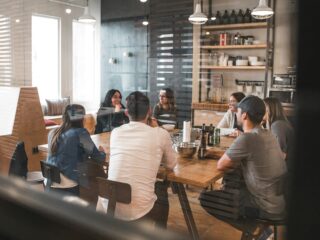 Group of people seated around a wooden table in a modern cafe, chatting and sharing drinks in a casual meeting.