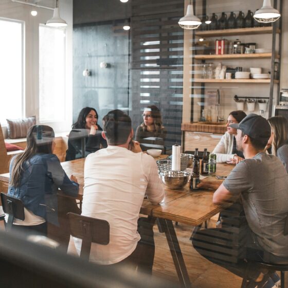 Group of people seated around a wooden table in a modern cafe, chatting and sharing drinks in a casual meeting.